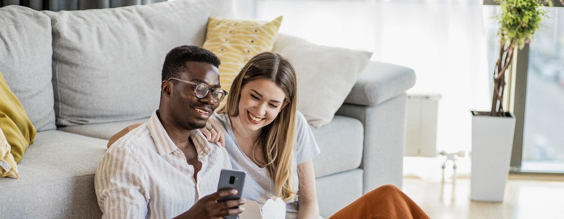 a man and woman with take out, sit against a couch on their living room floor and watch at their cell phone